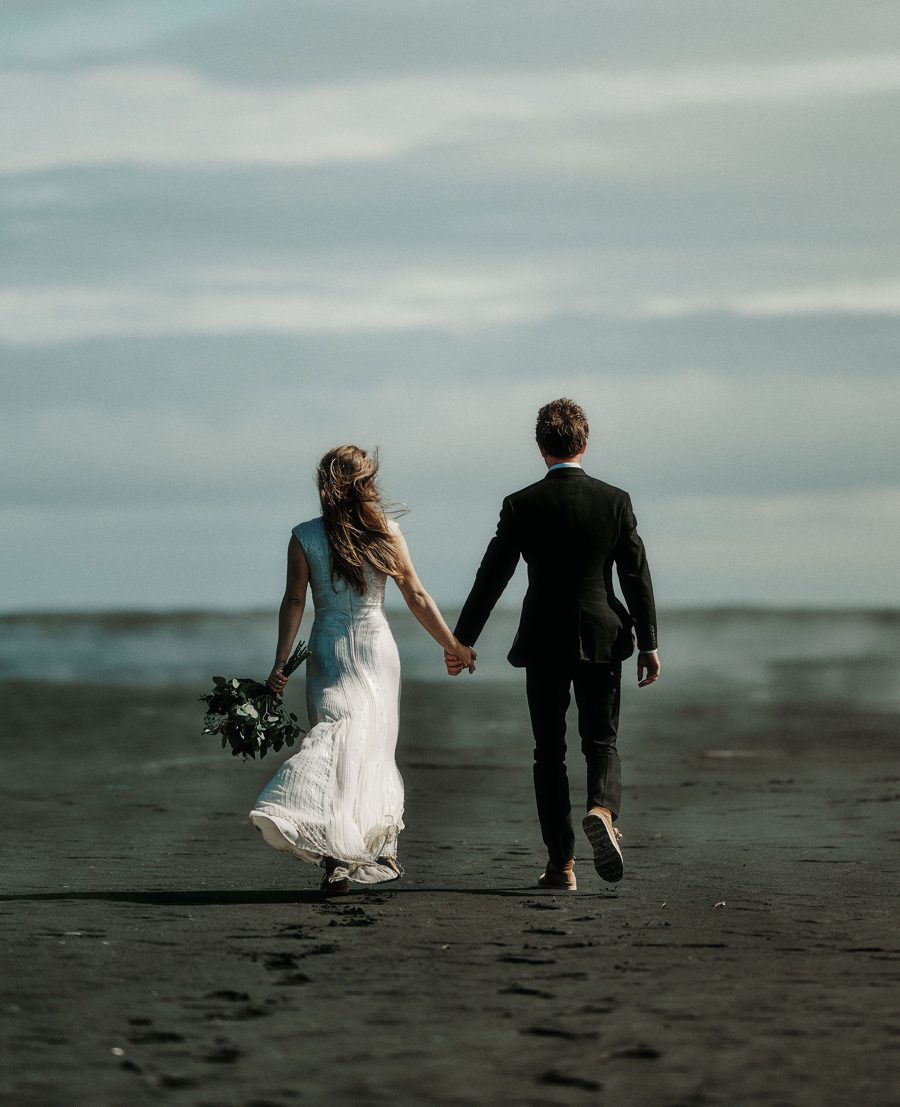 Matt Davis and Gareth Gwyn on their wedding day, seen from behind, walking hand in hand across a black sand beach toward the sea.